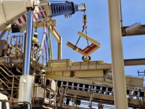 Construction workers use a crane to lift and position yellow structural beams during the assembly of an industrial facility.